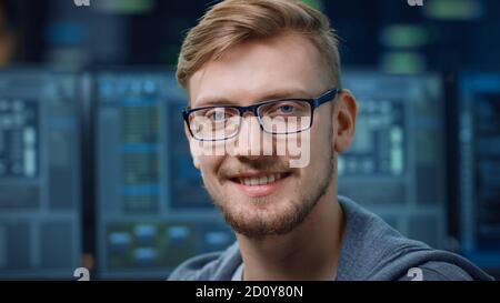 Portrait of a Smart and Handsome IT Specialist Wearing Glasses Smiles at the Camera. In the Background Unfocused Personal Computers with Screens in Stock Photo