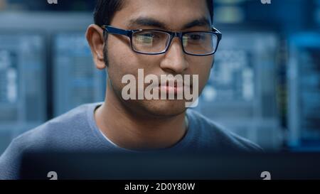 Portrait of a Smart and Handsome Software Developer Wearing Glasses Working on a Personal Computer, Smiles at the Camera. In the Background Unfocused Stock Photo