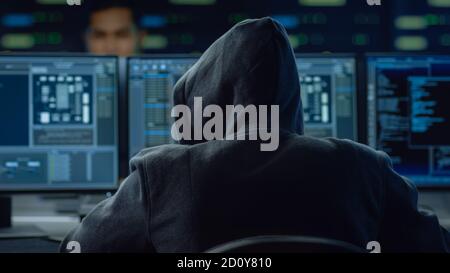 Hooded IT Hacker or Specialist Works on a Personal Computer With Display Showing Software Program with Coding Language Interface Stock Photo