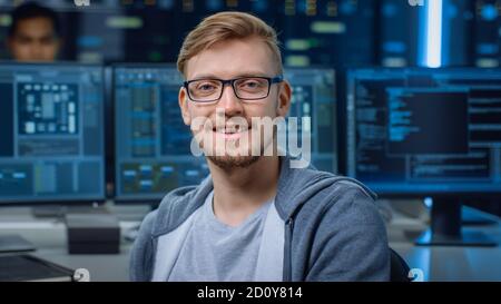 Portrait of a Smart and Handsome IT Specialist Wearing Glasses Smiles, Behind Him Personal Computers with Screens Showing Software Program with Coding Stock Photo