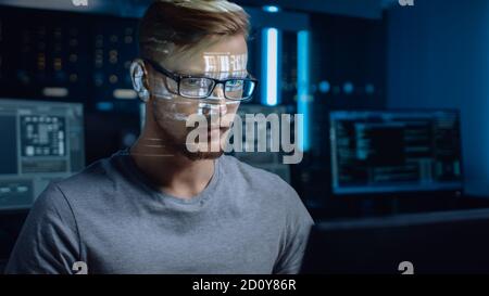 Portrait of Software Developer Hacker Working on Computer, Projected Code Numbers and Characters Reflect on His Face. Dark Room Full of Electronics Stock Photo