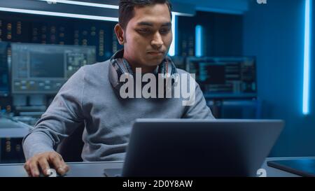 Portrait of Software Developer Hacker Programmer Sitting at His Desk and Working on Laptop Computer In the Background Dark Neon High Tech Environment Stock Photo