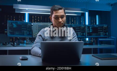 Portrait of Handsome Indian Software Developer Hacker Gamer Wearing Headset Sitting at His Desk and Working Playing on Laptop. In Background Dark Neon Stock Photo