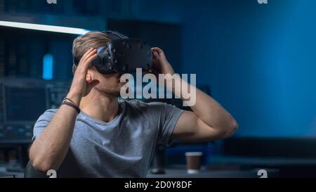 Software Delevoper Putting on Virtual Reality Headset on with His Head, Developing and Programming VR Game or Application. In the Background Stock Photo