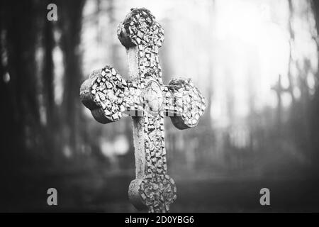 A black-and-white image of an old stone cracked cross standing in the middle of a foggy abandoned scary cemetery. Sadness. Death. Stock Photo