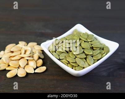 Bowl of tasty and healthy roasted pumpkin seeds with heap of raw seeds isolated on wooden table Stock Photo