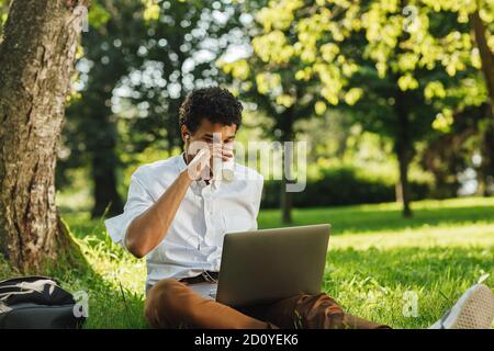 Young man sitting on the grass in park with laptop computer on his hips and drinking coffee Stock Photo