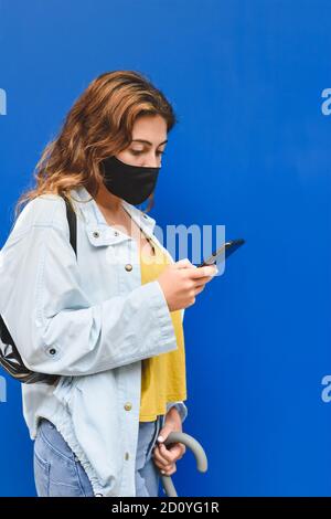 vertical portrait of a young redhead woman wearing yellow shirt standing in street using her phone and with blue wall background Stock Photo