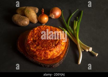 Potato omelette on a wooden board next to potatoes and onions on a black background Stock Photo