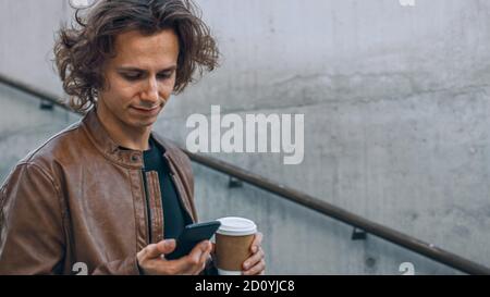Handsome Young Hispter Man with Long Hair Stands on the Stairs while Holding a Coffee and Looking at His Smartphone. He's in an Urban Environment.  Stock Photo