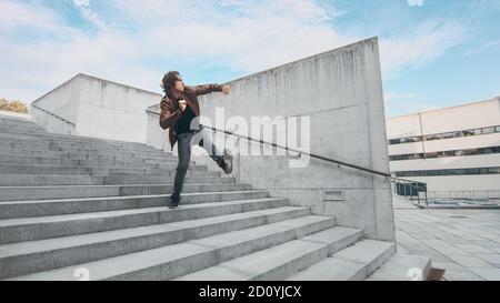 Cheerful and Happy Young Man with Long Hair Actively Dancing While Walking Down the Stairs. He's Wearing a Brown Leather Jacket. Scene Shot in an Stock Photo