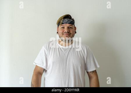 A smiling Caucasian male with tattoos wearing a bandana on his head against a wall Stock Photo