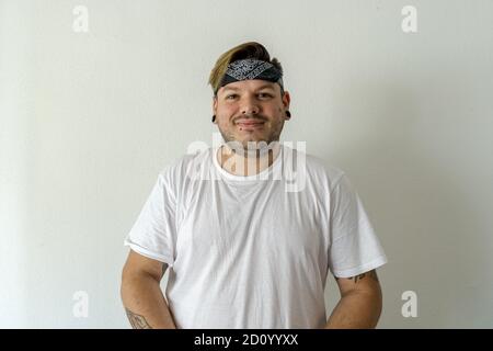 A smiling Caucasian male with tattoos wearing a bandana on his head against a wall Stock Photo