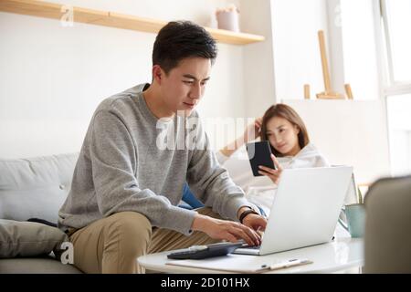 young asian couple staying at home with husband working using laptop computer and wife  reading ebook Stock Photo