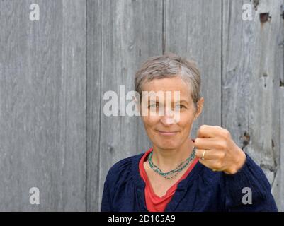 Serious middle-aged Caucasian country woman with short hair threatens the viewer with her clenched left fist, in front of old barn wood background. Stock Photo