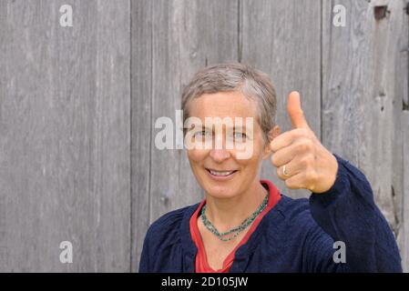 Smiling middle-aged Caucasian country woman shows thump up (approval sign) with ambiguous emotions, in front of an old barn wood background. Stock Photo