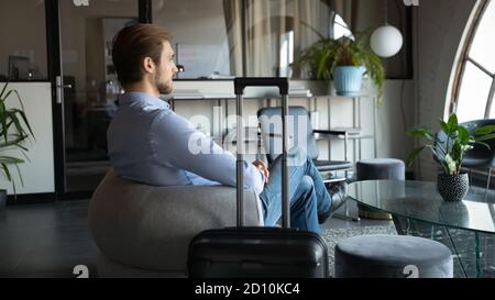 Side view pensive young man sitting with luggage at office. Stock Photo