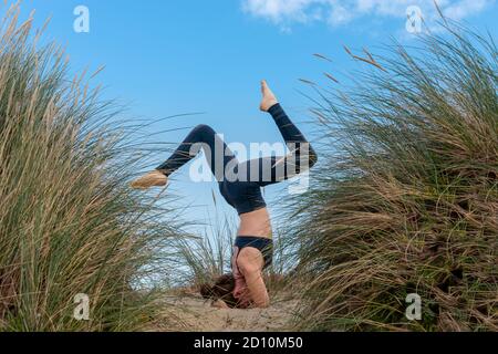 Woman doing a headstand in sand dunes with blue sky background. Yoga and exercise outdoors. Stock Photo
