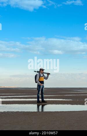 Man carrying a tripod standing on sand by the coast. Photographer with equipment. Stock Photo