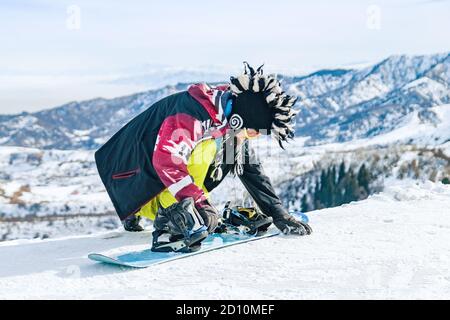 Young man snowboarder in fanny hat sits in the snow and fastens fastenings on a snowboard before the descent Stock Photo