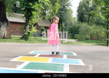 Little girl in a pink dress playing hopscotch on playground outdoors, children outdoor activities Stock Photo