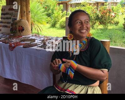 Nueva Loja, Sucumbios / Ecuador - September 2 2020: Indigenous woman of Cofan nationality with green dress smiling while weaving handicrafts sitting o Stock Photo