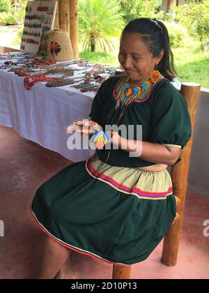Nueva Loja, Sucumbios / Ecuador - September 2 2020: Indigenous woman of Cofan nationality with green dress smiling while weaving handicrafts sitting o Stock Photo