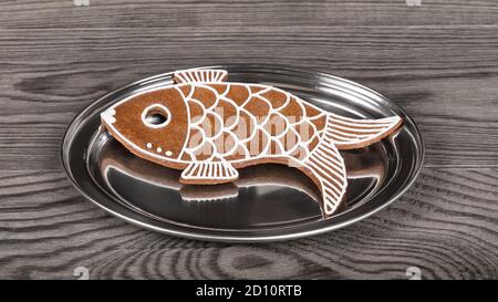 Sweet fish shaped gingerbread cookie with reflection on round silver plate. Xmas pastry decorated with white icing on shiny stainless serving salver. Stock Photo