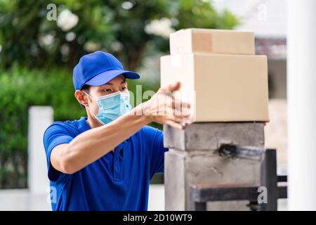 Asian deliver man with face mask in blue shirt handling packages and put in on the fence post of customer house as contactless shopping delivery. This Stock Photo