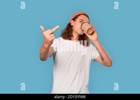 Freestyle. Curly-haired guy wearing hat standing isolated on blue drinking coffee showing middle finger cool Stock Photo