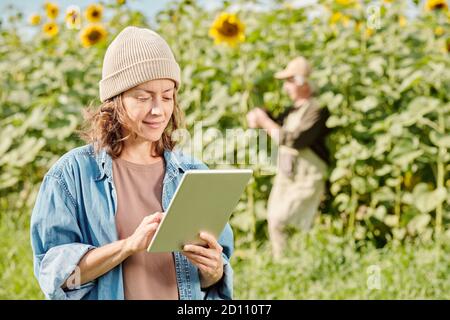 Mature female farmer in workwear standing in front of camera and using touchpad Stock Photo