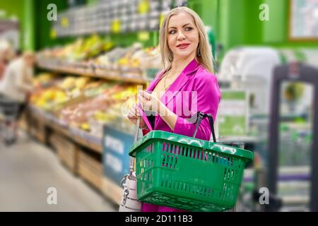 Mature woman in pink inside a supermarket with a green shopping basket. A middle-aged white smiling female purchases food from a grocery store. Stock Photo