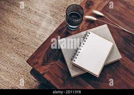 Empty diary, notepad mockup and book on wooden table. Glass of water and dry Lagurus ovatus grass. Blurred beige jute carpet background. Flat lay, top Stock Photo