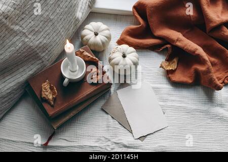 Autumn still life. Burning candle, books and linen pillow near window. Blank greeting card mockup scene. Moody composition with leaves and white Stock Photo