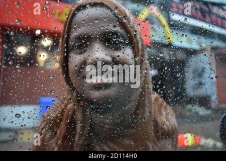 DHAKA, BANGLADESH- AUGUST 14, 2020 : Portrait of female Bangladeshi poor kid while smiling with poverty face in a rainy day at road, water drops backg Stock Photo
