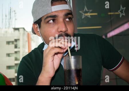 Mustaches young man with mask and cap in new normal of CORONAVIRUS drinking with straw and looking with eyes in a restaurant, polo green shirt and eat Stock Photo