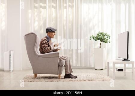 Elderly man with a cup of tea sitting in an armchair and watching tv Stock Photo