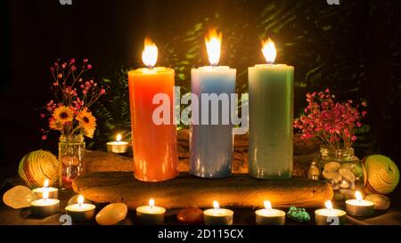 Composition with three large green, red and blue candles surrounded by white candles, flowers and stones on a black background with green light Stock Photo