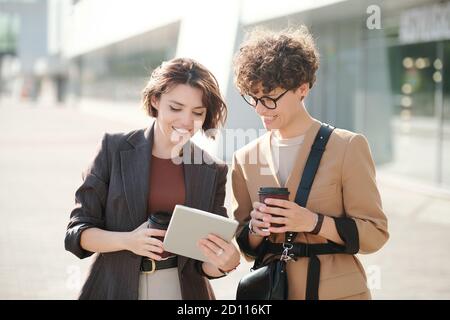 Two happy young colleagues with drinks looking at screen of digital tablet Stock Photo