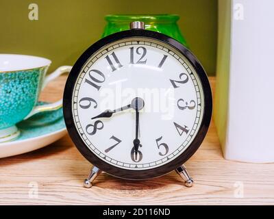 An alarm clock with a black case and black numbers and arrows on a white dial stands on a wooden table Stock Photo