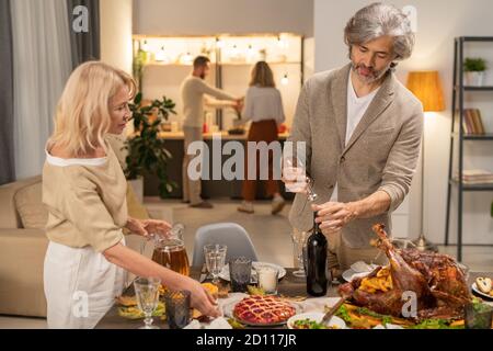 Serious mature bearded man opening bottle of wine while standing by his wife Stock Photo