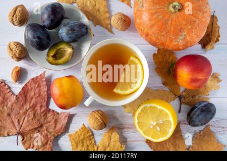 Autumn background of dry leaves with cup of tea, a small beautiful pumpkin, lemon, ripe plums and nectarines, walnuts. Stock Photo