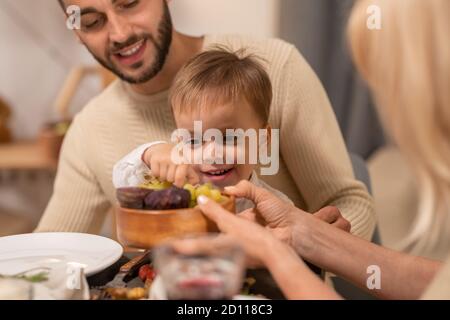 Happy young man with cute little son on his knees looking at him taking grape Stock Photo