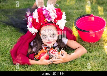 Cute girl with painted face holding pile of sweet halloween treats on green lawn Stock Photo