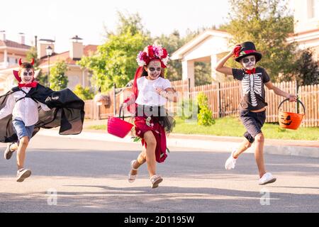 Three laughing kids in halloween costumes running down wide road on sunny day Stock Photo