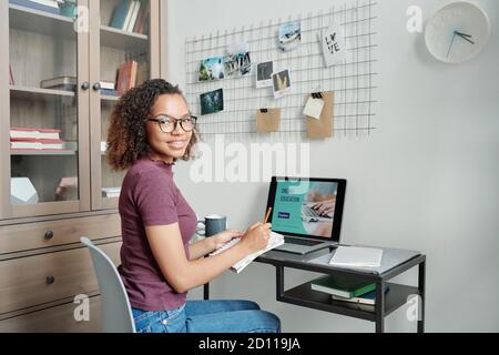 Young cheerful female student in casualwear making notes in front of laptop Stock Photo