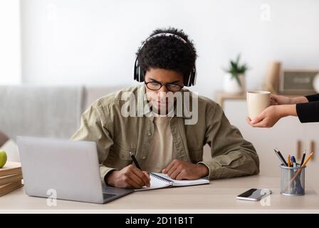 Black teen guy in headphones studying online from home, making notes in copybook Stock Photo