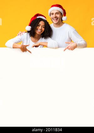 Happy Couple Wearing Santa Hats And Pointing At White Advertisement Board Stock Photo
