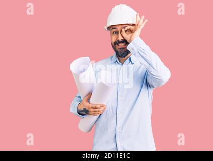 Young handsome man wearing hardhat holding paper blueprints smiling happy doing ok sign with hand on eye looking through fingers Stock Photo