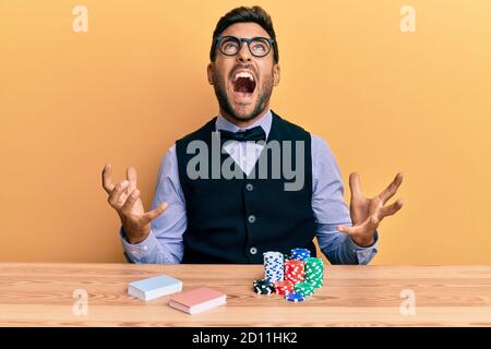 Handsome hispanic croupier man sitting on the table with poker chips and cards crazy and mad shouting and yelling with aggressive expression and arms Stock Photo